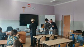 W sali lekcyjnej z fioletowymi ścianami i krzyżem nad tablicą multimedialną dwoje uczniów i instruktor obserwują ekran komputera. Inni uczniowie siedzą w ławkach, a instruktor trzyma kontrolery, a jeden z uczniów ma na sobie gogle VR.