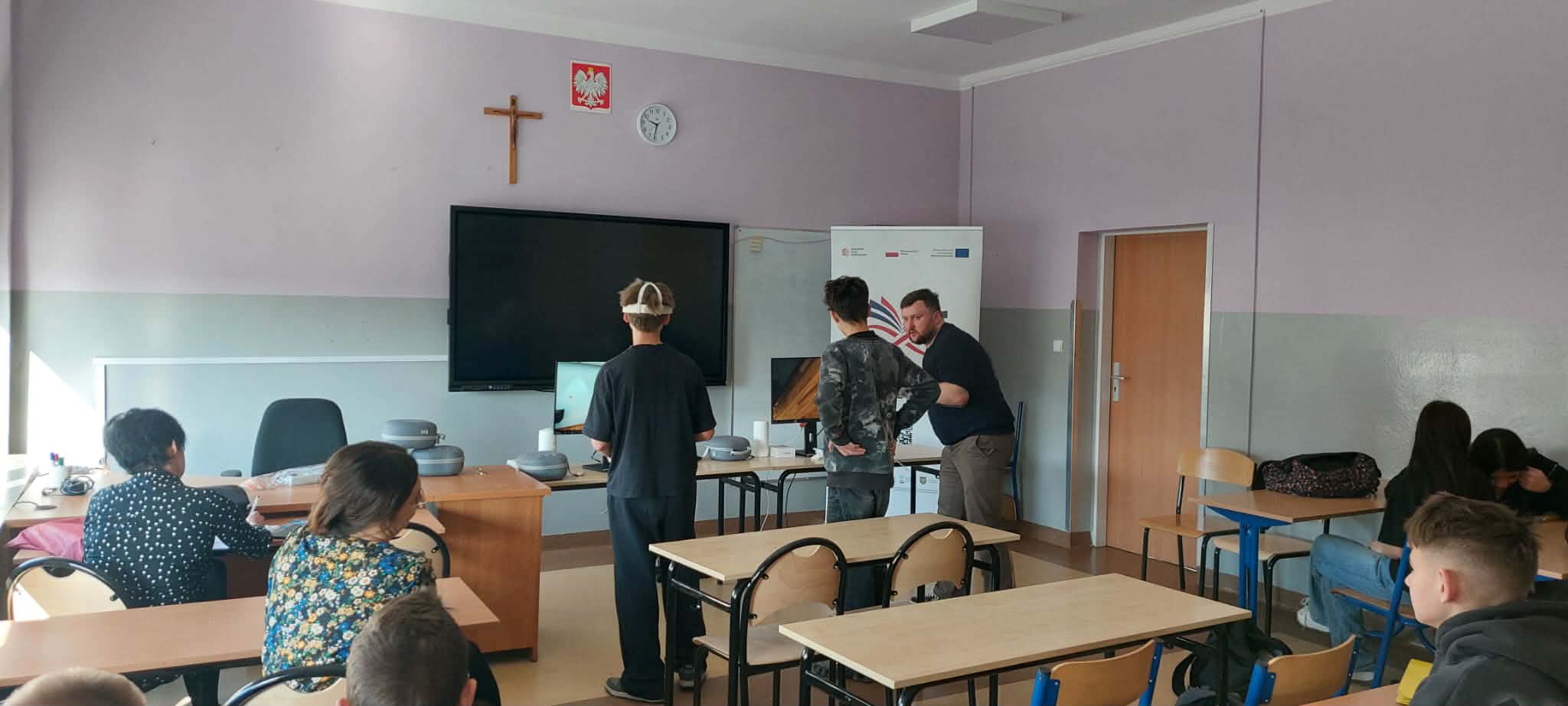 W sali lekcyjnej z fioletowymi ścianami i krzyżem nad tablicą multimedialną dwoje uczniów i instruktor obserwują ekran komputera. Inni uczniowie siedzą w ławkach, a instruktor trzyma kontrolery, a jeden z uczniów ma na sobie gogle VR.