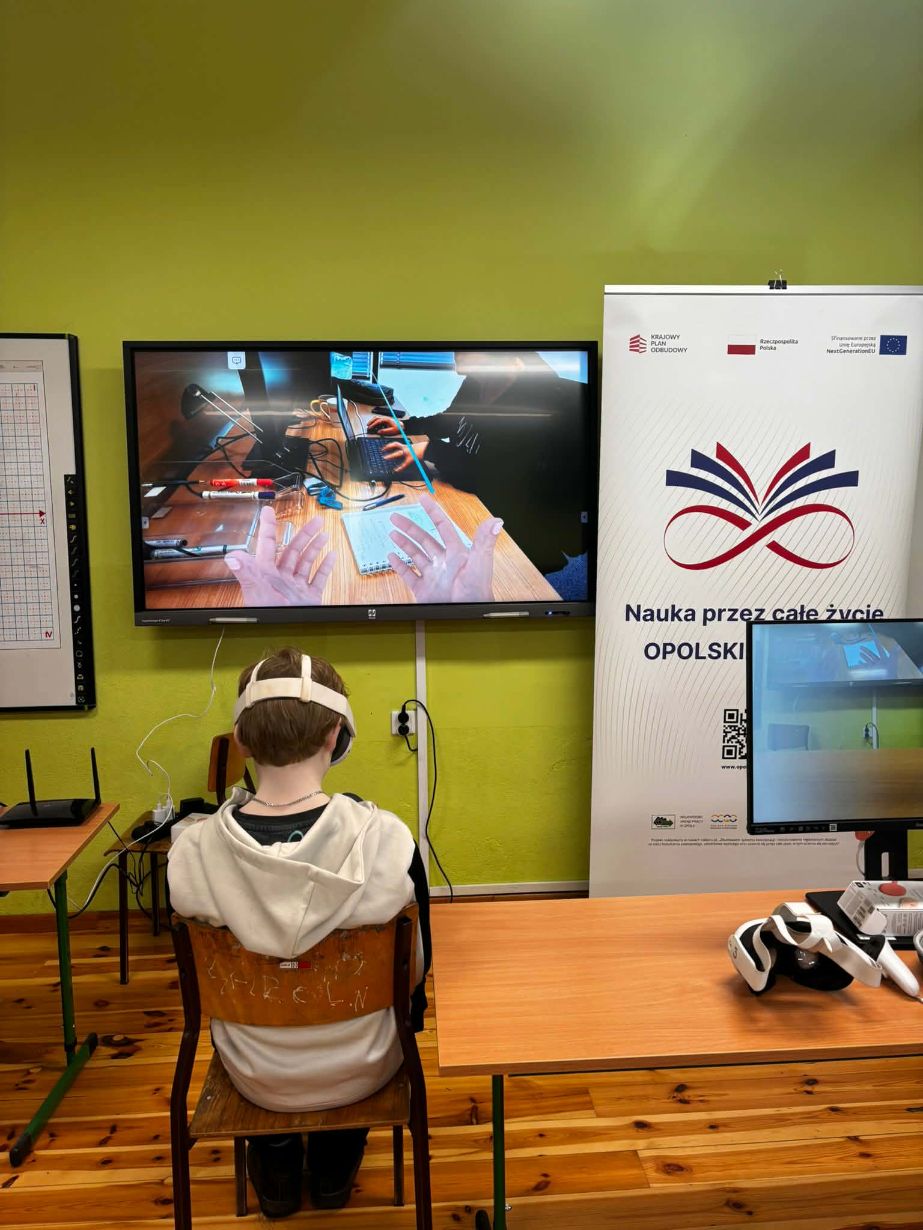 Zbliżenie na ucznia siedzącego tyłem do aparatu w białych goglach VR, który patrzy na duży interaktywny ekran. Na wyświetlaczu widać widok z perspektywy pierwszej osoby, przedstawiający ręce w wirtualnej rzeczywistości operujące nad biurkiem z laptopem i notatkami. Po prawej stronie stoi biały baner informacyjny z logotypami Unii Europejskiej i programów regionalnych. Na stole obok leży dodatkowy zestaw gogli i akcesoria.