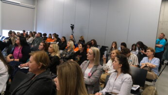 Ujęcie z boku na uczestników konferencji wypełniających salę. Ludzie siedzą na krzesłach, niektórzy robią zdjęcia telefonami lub notują. Jasne ściany i industrialny sufit nadają wnętrzu surowy, lecz nowoczesny wygląd.