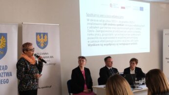 Prelegentka w kolorowej bluzce i okularach przemawia do mikrofonu podczas konferencji. Obok niej przy stole siedzą trzy osoby, w tym mężczyzna w garniturze i kobieta w różowej bluzce. W tle widoczny jest ekran z prezentacją zatytułowaną „Spotkania sieciująco-edukacyjne”.