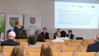 Szerokie ujęcie sali konferencyjnej wypełnionej słuchaczami siedzącymi w rzędach krzeseł. Na przedzie kobieta z krótkimi włosami prowadzi prezentację przy ekranie z logotypami unijnymi. Obok niej znajdują się banery informacyjne z godłem województwa opolskiego.