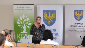 Zbliżenie na kobietę w okularach i pomarańczowym szalu, która trzyma mikrofon i pilot do slajdów. Stoi ona na tle zielonego baneru Regionalnego Centrum Rozwoju Edukacji w Opolu oraz herbu województwa. Przed nią widoczne są tyły głów uczestników spotkania.