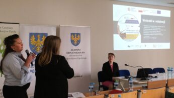 Kobieta z ciemnymi upiętymi włosami przemawia do mikrofonu, gestykulując dłońmi. W tle widać fragment ekranu z grafiką „Wnioski w edukacji” oraz siedzącą przy stole inną uczestniczkę. Na pierwszym planie znajdują się butelki wody i materiały konferencyjne.