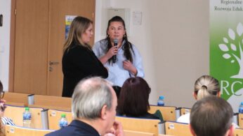 Dwie kobiety rozmawiają ze sobą na środku sali; jedna z nich trzyma mikrofon i patrzy w stronę rozmówczyni. W dolnej części kadru widać uczestników siedzących tyłem, słuchających wystąpienia. W tle znajdują się jasne ściany i drewniane drzwi.