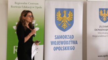Portret prelegentki w czarnej sukience i okularach, która mówi do mikrofonu, trzymając w drugiej ręce kartkę z notatkami. Stoi ona pomiędzy zielonym banerem edukacyjnym a białym banerem Samorządu Województwa Opolskiego.