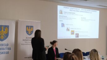 Prelegentka tyłem do obiektywu wskazuje na ekran z wyświetloną prezentacją dotyczącą systemu kształcenia w powiecie krapkowickim. Przy stole konferencyjnym siedzi inna kobieta w różowej bluzce, uważnie patrząc w stronę widowni