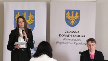 Kobieta w czarnej marynarce i białym golfie przemawia do mikrofonu, patrząc przed siebie. Stoi bezpośrednio przed banerem z napisem „Zuzanna Donath-Kasiura, Wicemarszałek Województwa Opolskiego”. Obok niej przy laptopie siedzi inna uczestniczka wydarzenia