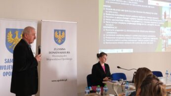 Starszy mężczyzna w ciemnej marynarce przemawia do mikrofonu, stojąc z boku sali. Na ekranie za nim wyświetlane są wnioski i rekomendacje dotyczące uczenia się przez całe życie. W tle widoczne są banery instytucjonalne oraz siedząca przy stole kobieta.