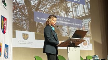 Kobieta w ciemnej marynarce stoi przy mównicy na tle dużego ekranu projekcyjnego. Na slajdzie widnieje hasło „Z PASJĄ DO POMAGANIA, Z PEWNOŚCIĄ ZATRUDNIENIA” oraz zdjęcie budynku. Po lewej stronie sceny ustawiona jest ścianka z herbami miast województwa opolskiego.