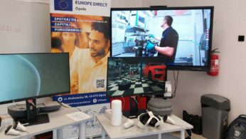 Zbliżenie na stanowisko technologiczne. Na białym stole leżą dwa zestawy gogli VR z kontrolerami oraz szare etui. Nad stołem znajdują się dwa monitory: lewy wyświetla błękitną grafikę sferyczną, prawy pokazuje wnętrze wirtualnego warsztatu samochodowego z czerwonym autem. Obok, na ścianie, zamontowany jest duży telewizor wyświetlający film z pracownikiem technicznym przy maszynie. W tle baner Europe Direct Opole.