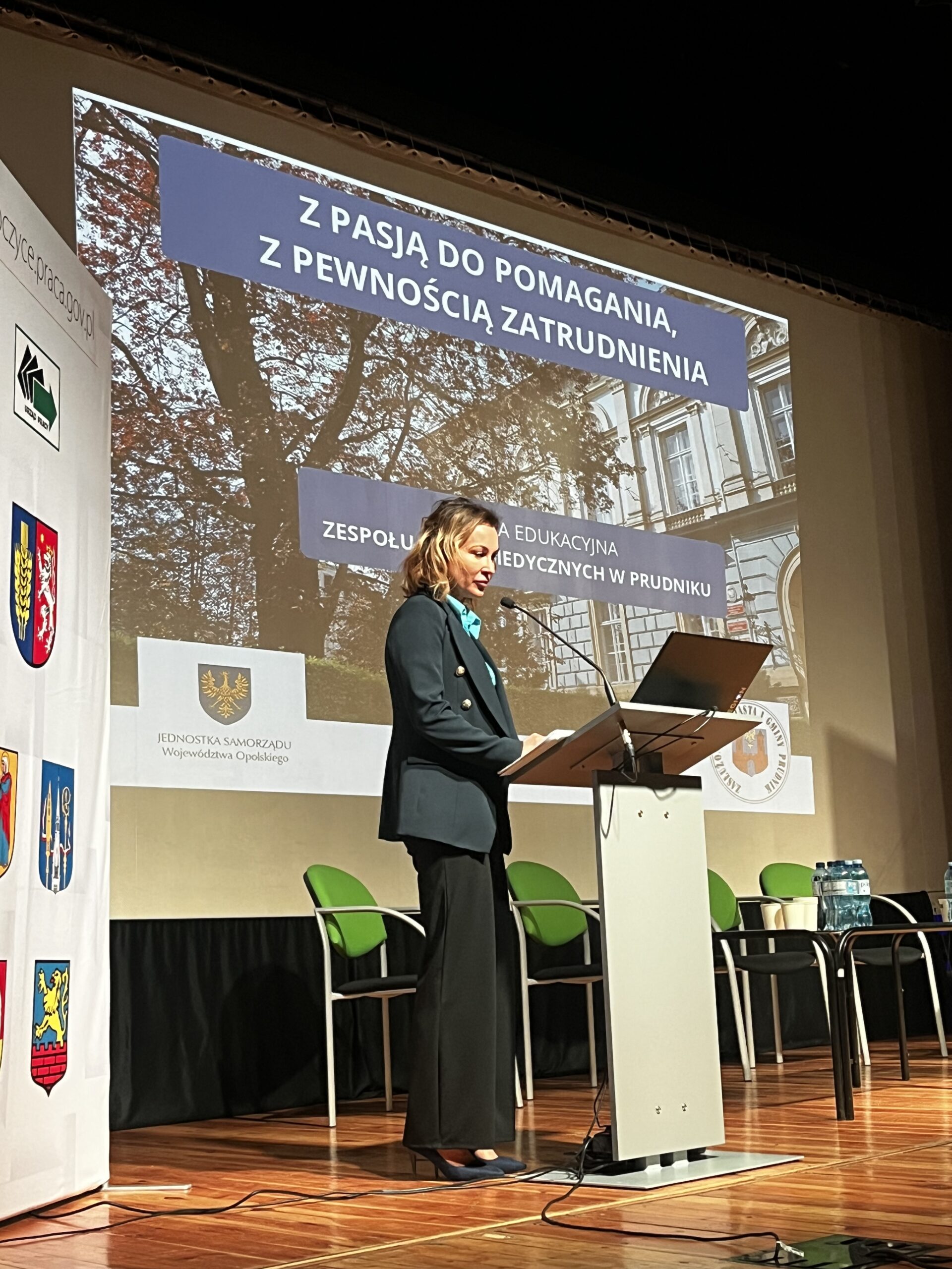 Kobieta w ciemnej marynarce stoi przy mównicy na tle dużego ekranu projekcyjnego. Na slajdzie widnieje hasło „Z PASJĄ DO POMAGANIA, Z PEWNOŚCIĄ ZATRUDNIENIA” oraz zdjęcie budynku. Po lewej stronie sceny ustawiona jest ścianka z herbami miast województwa opolskiego.