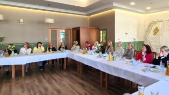 Ujęcie rzędu uczestniczek konferencji siedzących przy wspólnym stole przykrytym białym obrusem, na którym stoją napoje i przekąski. Kobiety z uwagą patrzą przed siebie, a w tle widać eleganckie wnętrze lokalu z drewnianymi drzwiami i dekoracyjną ścianką.