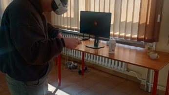 Uczeń w ciemnej bluzie stoi w nasłonecznionej sali, korzystając z gogli VR i kontrolerów ruchu. Na biurku obok niego znajduje się monitor oraz akcesoria higieniczne do dezynfekcji sprzętu. Przez żaluzje w oknach wpada jasne światło, tworząc wyraźne cienie na podłodze.