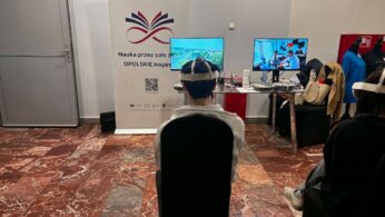 Widok zza pleców siedzącej osoby w goglach VR, która patrzy w stronę dwóch monitorów wyświetlających różne sceny wideo. Po lewej stronie stoi duży baner z napisem „Nauka przez całe życie”. Na szklanym stole leżą pokrowce na sprzęt elektroniczny i materiały informacyjne.