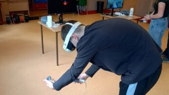 Osoba w czarnej bluzie i spodniach dresowych pochyla się nisko, trzymając kontrolery VR podczas sesji wirtualnej rzeczywistości. W tle widać drugą osobę w goglach oraz stanowiska z monitorami i sprzętem elektronicznym. Sala jest przestronna, z wyraźnie widocznym ekranem projekcyjnym w głębi.