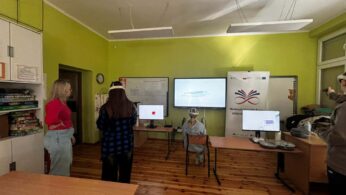 Szerokie ujęcie sali lekcyjnej, w której grupa osób testuje technologię rzeczywistości wirtualnej. Trzy osoby mają na głowach gogle VR i są zwrócone w stronę monitorów oraz tablicy interaktywnej wyświetlającej prezentację. Po lewej stronie stoi kobieta w czerwonej bluzce, nadzorująca przebieg zajęć. W pomieszczeniu znajdują się drewniane stoły, krzesła oraz regał wypełniony grami planszowymi i materiałami edukacyjnymi.