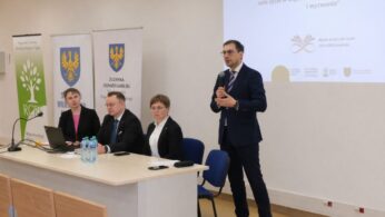 Zdjęcie przedstawia podium z profilu, na którym stoi mężczyzna przemawiający do mikrofonu. Przy długim stole siedzą dwie kobiety i jeden mężczyzna, a przed nimi stoją butelki wody i laptop. W tle widoczne są flagi i banery informacyjne, w tym tablica z napisem 