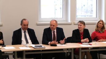 Czworo uczestników konferencji siedzi przy wspólnym stole na tle białej ściany z oknami. Dwaj mężczyźni w środkowej części zdjęcia aktywnie gestykulują i rozmawiają, podczas gdy dwie kobiety po bokach przysłuchują się debacie. Na blacie leżą teczki z dokumentami, notatniki oraz filiżanka.