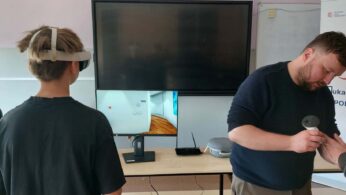 W klasie uczeń w goglach VR stoi przed ekranem komputera, a instruktor pomaga mu z kontrolerami. Na stole leżą gogle, kontrolery i router, a w tle widać baner z logo i napisem 