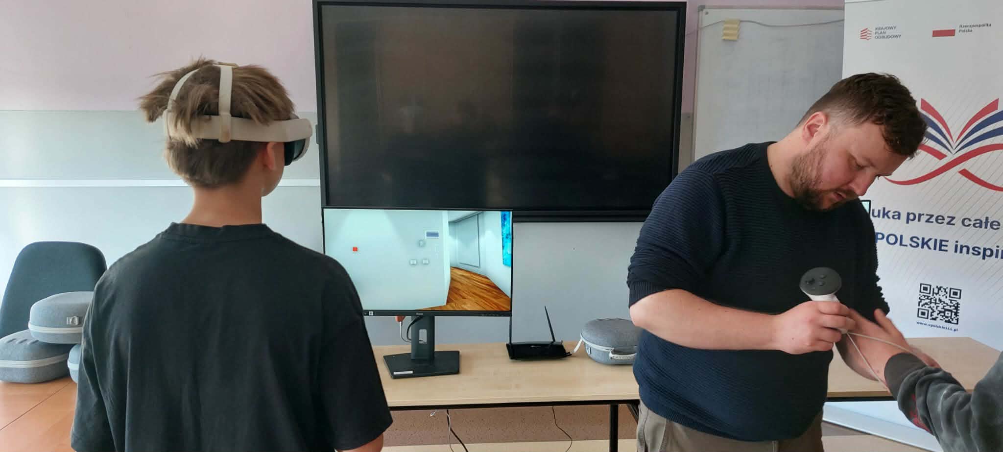 W klasie uczeń w goglach VR stoi przed ekranem komputera, a instruktor pomaga mu z kontrolerami. Na stole leżą gogle, kontrolery i router, a w tle widać baner z logo i napisem 