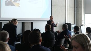 Widok zza pleców publiczności na mężczyznę przemawiającego do mikrofonu podczas oficjalnej konferencji. W tle znajduje się duży ekran projekcyjny z napisem dotyczącym wsparcia nowoczesnego kształcenia zawodowego w Opolu, z datą 11.03.2026. Uczestnicy siedzą w rzędach, a niektórzy z nich klaszczą.