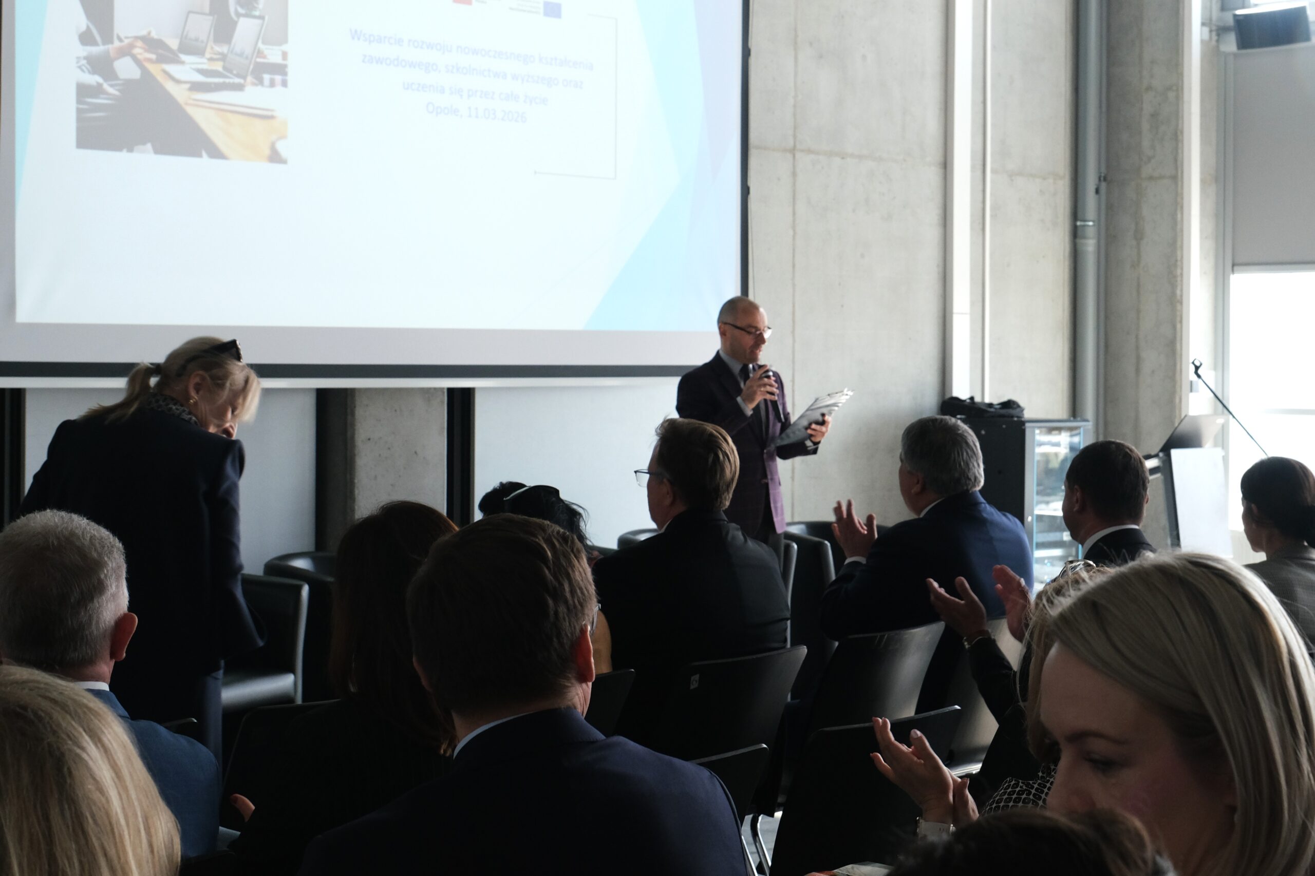 Widok zza pleców publiczności na mężczyznę przemawiającego do mikrofonu podczas oficjalnej konferencji. W tle znajduje się duży ekran projekcyjny z napisem dotyczącym wsparcia nowoczesnego kształcenia zawodowego w Opolu, z datą 11.03.2026. Uczestnicy siedzą w rzędach, a niektórzy z nich klaszczą.
