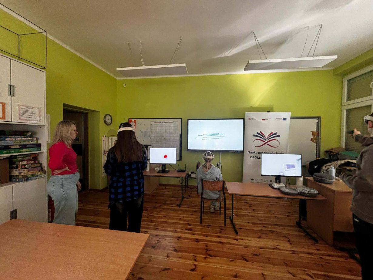 Szerokie ujęcie sali lekcyjnej, w której grupa osób testuje technologię rzeczywistości wirtualnej. Trzy osoby mają na głowach gogle VR i są zwrócone w stronę monitorów oraz tablicy interaktywnej wyświetlającej prezentację. Po lewej stronie stoi kobieta w czerwonej bluzce, nadzorująca przebieg zajęć. W pomieszczeniu znajdują się drewniane stoły, krzesła oraz regał wypełniony grami planszowymi i materiałami edukacyjnymi.