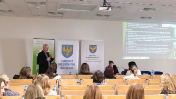 Szerokie ujęcie sali konferencyjnej z rzędami krzeseł, w której trwa prelekcja. Starszy mężczyzna w ciemnej marynarce przemawia do mikrofonu, stojąc obok banerów Województwa Opolskiego. W tle widoczny jest ekran projekcyjny z wyświetloną prezentacją na temat uczenia się przez całe życie.