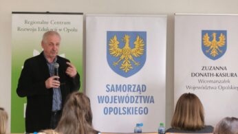 Zbliżenie na starszego mężczyznę przemawiającego do mikrofonu podczas konferencji. Prelegent stoi na tle białych banerów z herbem województwa opolskiego i napisem „Samorząd Województwa Opolskiego”. W dolnej części kadru widać głowy uczestników siedzących w ławkach.