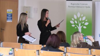 Dwie kobiety stojące w sali konferencyjnej, z których jedna przemawia do mikrofonu, trzymając w dłoniach dokumenty. W tle widoczny jest zielony baner Regionalnego Centrum Rozwoju Edukacji w Opolu (RCRE). Na pierwszym planie siedzą uczestnicy spotkania, a w tle widać drzwi z naklejonym plakatem.