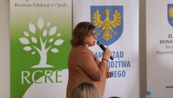 Profilowe ujęcie kobiety w jasnobrązowej marynarce i okularach, która przemawia do mikrofonu. Postać znajduje się na tle zielonego baneru z logo drzewa oraz białego baneru z herbem. W dolnej części zdjęcia widoczne są rozmyte sylwetki słuchaczy.