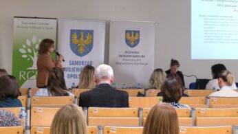 Widok z perspektywy uczestnika na salę konferencyjną, gdzie kobieta w brązowym żakiecie prowadzi prezentację. Obok niej znajdują się oficjalne banery informacyjne, a po prawej stronie widać fragment slajdu z wnioskami i rekomendacjami. W rzędach przed prelegentką siedzą tyłem do obiektywu uczestnicy spotkania.