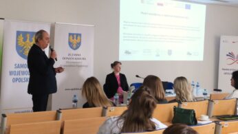 Mężczyzna w ciemnym garniturze przemawia do mikrofonu, stojąc przed ekranem z prezentacją zatytułowaną „Model współpracy w obszarze doradztwa i LLL”. Po prawej stronie przy stole siedzi kobieta w różowej bluzce, pełniąca rolę panelistki lub protokolantki. Na pierwszym planie widoczne są plecy uczestników siedzących w sali.