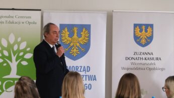 Portretowe ujęcie mężczyzny w garniturze przemawiającego podczas oficjalnego spotkania. W tle znajdują się dwa białe banery z herbem województwa opolskiego, w tym jeden z nazwiskiem Zuzanny Donath-Kasiury. Atmosfera zdjęcia sugeruje formalny charakter wydarzenia regionalnego.