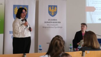 Kobieta w białej koszuli ozdobionej motywami kart do gry i w okularach przemawia do mikrofonu. Stoi obok banerów promocyjnych województwa, a w tle widoczna jest siedząca przy stole kolejna uczestniczka. Na pierwszym planie widać tył głowy jednej z osób siedzących w auli.