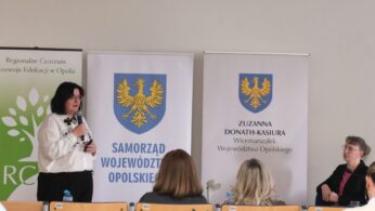 Ujęcie prelegentki w białej bluzce z mikrofonem w ręku, wykonane podczas wystąpienia w sali wykładowej. Kobieta stoi obok baneru z napisem „Samorząd Województwa Opolskiego”. Po prawej stronie kadru siedzi inna kobieta w czarnej marynarce, obserwująca przebieg spotkania.