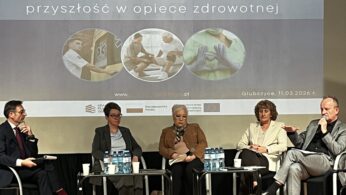 Grupa pięciu osób siedzi w fotelach na scenie podczas panelu dyskusyjnego. Nad nimi wyświetlany jest napis „ZAWODY ODPORNE NA KRYZYS - przyszłość w opiece zdrowotnej” wraz z datą 11.03.2026 r. Na stolikach przed panelistami stoją butelki z wodą i papierowe kubki.