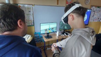 Zbliżenie na ucznia w okularach VR, który za pomocą kontrolerów manipuluje obiektami w cyfrowym warsztacie. Na monitorze widać wirtualne puszki z farbą oraz interfejs aplikacji do nauki zawodu. Obok stoi opiekun lub nauczyciel, który obserwuje postępy na ekranie. Scena pokazuje praktyczne wykorzystanie technologii w edukacji.