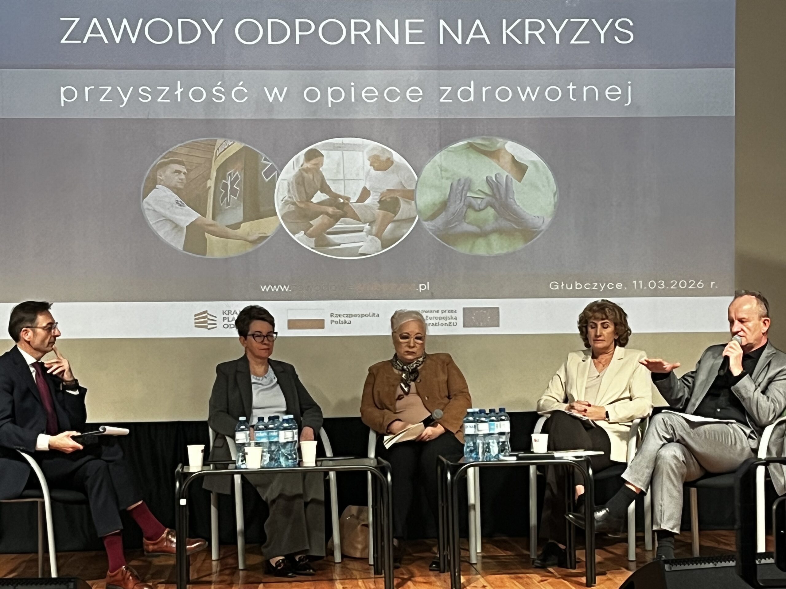 Grupa pięciu osób siedzi w fotelach na scenie podczas panelu dyskusyjnego. Nad nimi wyświetlany jest napis „ZAWODY ODPORNE NA KRYZYS - przyszłość w opiece zdrowotnej” wraz z datą 11.03.2026 r. Na stolikach przed panelistami stoją butelki z wodą i papierowe kubki.