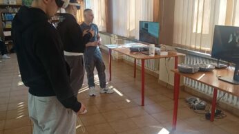 Dwóch uczniów w goglach VR stoi wzdłuż rzędu biurek w jasnej sali, podczas gdy instruktor nadzoruje ich działania. Na stolikach rozstawione są monitory pomocnicze oraz etui na zestawy wirtualnej rzeczywistości. W głębi pomieszczenia widoczne są regały z książkami.