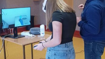 Zbliżenie na osobę w goglach VR i niebieskich jeansach, która trzyma kontroler przed monitorem. Obok niej stoi mężczyzna w niebieskiej bluzie z kapturem, udzielając wskazówek lub obserwując postępy. Monitor na biurku pokazuje widok wnętrza z perspektywy pierwszej osoby w aplikacji VR.