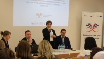 Kobieta w okularach i czarnym żakiecie stoi na podium i przemawia do mikrofonu, gestykulując dłonią. Po jej bokach siedzą pozostali uczestnicy panelu, a w tle widać ekran z prezentacją. Na pierwszym planie widać rozmazane sylwetki osób słuchających debaty.
