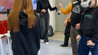 Grupa osób w jasnej sali lekcyjnej podczas warsztatów. Dwie dziewczyny mają na głowach białe gogle VR i trzymają kontrolery. Obok nich stoi dwóch instruktorów, którzy pomagają w obsłudze sprzętu. Nad drzwiami widoczny jest zegar oraz zielony znak „Wyjście ewakuacyjne”.