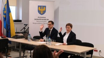 Dwoje prelegentów siedzi przy stole konferencyjnym, unosząc dłonie w geście powitania lub podczas głosowania. Przed nimi stoją butelki wody mineralnej, a w tle widoczna jest flaga oraz baner informacyjny. Na pierwszym planie widać zarys głowy innej osoby uczestniczącej w spotkaniu.