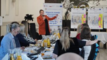 Kobieta w pomarańczowej marynarce stoi przed grupą słuchaczy, trzymając w górze dokument i przemawiając do mikrofonu. Za nią znajdują się plakaty informacyjne z kolorowymi karteczkami samoprzylepnymi, sugerującymi metodę pracy warsztatowej. Uczestnicy siedzący tyłem do obiektywu skupiają uwagę na prelegentce. Scena dokumentuje aktywną część prezentacji projektu.