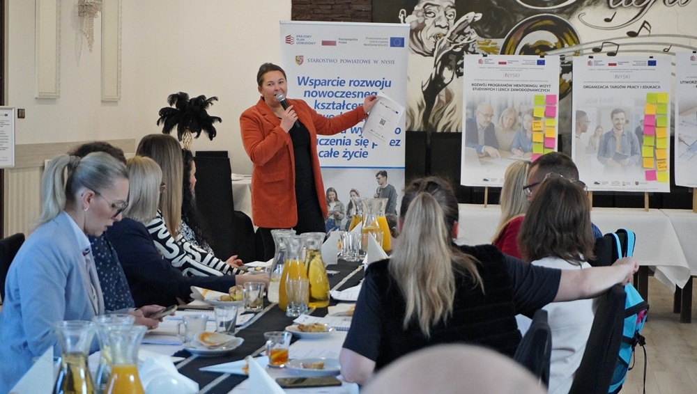 Kobieta w pomarańczowej marynarce stoi przed grupą słuchaczy, trzymając w górze dokument i przemawiając do mikrofonu. Za nią znajdują się plakaty informacyjne z kolorowymi karteczkami samoprzylepnymi, sugerującymi metodę pracy warsztatowej. Uczestnicy siedzący tyłem do obiektywu skupiają uwagę na prelegentce. Scena dokumentuje aktywną część prezentacji projektu.