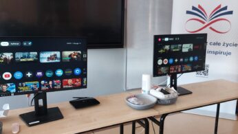Na drewnianym stole w klasie stoją dwa monitory wyświetlające menu oraz zestaw gogli VR z kontrolerami. Obok monitorów znajduje się czarny router i pilot, a w tle widać baner promocyjny.