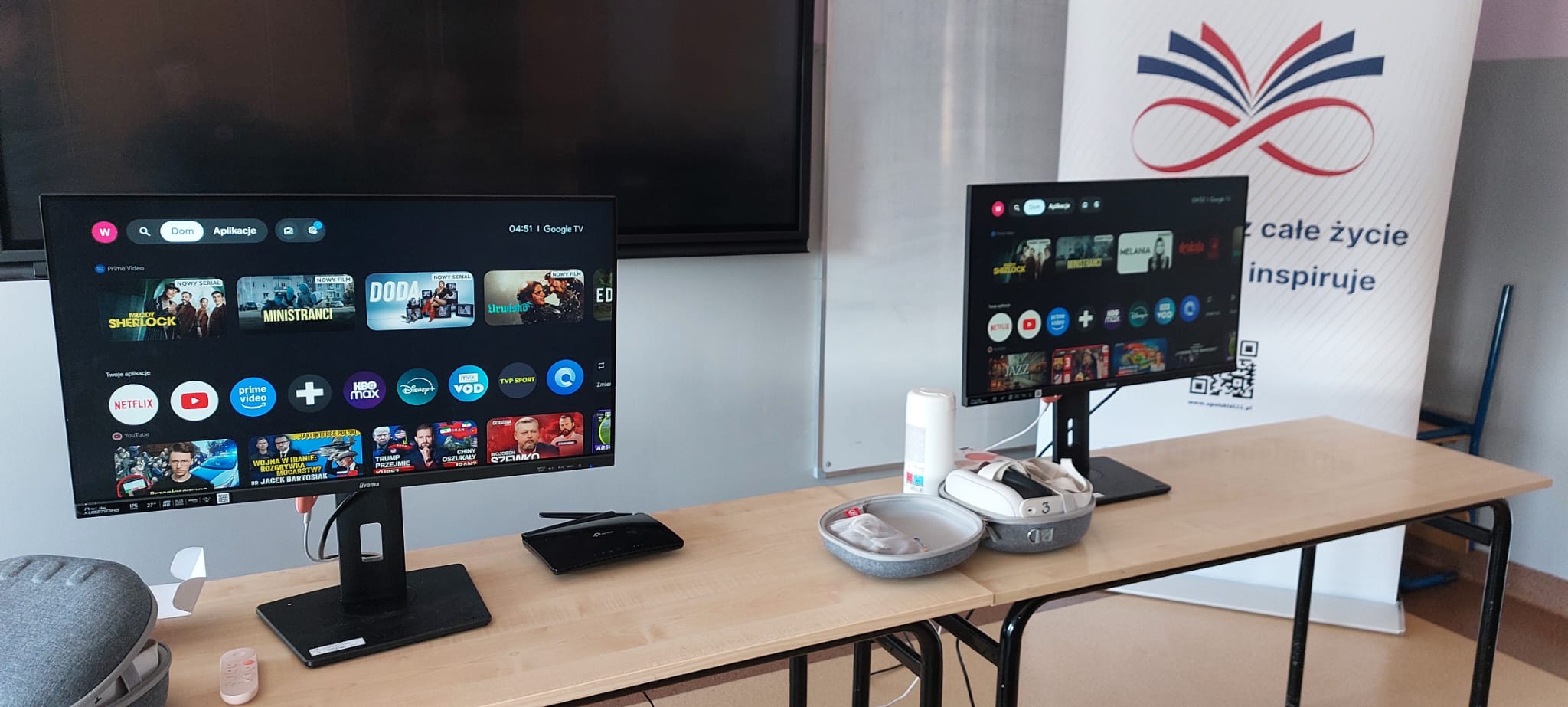 Na drewnianym stole w klasie stoją dwa monitory wyświetlające menu oraz zestaw gogli VR z kontrolerami. Obok monitorów znajduje się czarny router i pilot, a w tle widać baner promocyjny.