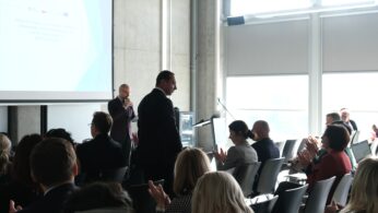 Szerokie ujęcie sali konferencyjnej, na którym widać profil mężczyzny w garniturze stojącego przed siedzącą publicznością. W tle, przy ekranie, kolejna osoba przemawia do mikrofonu, a zgromadzeni goście reagują brawami. Sala jest jasna, z dużymi oknami i surowymi, betonowymi ścianami.